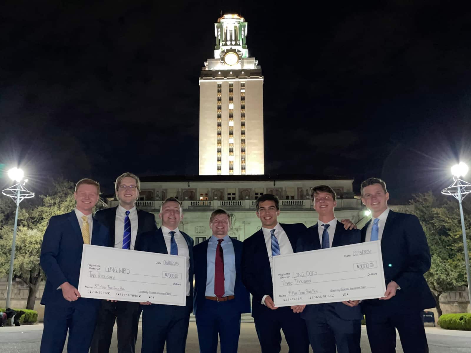 Seven students hold two large checks from a stock competition outside at night