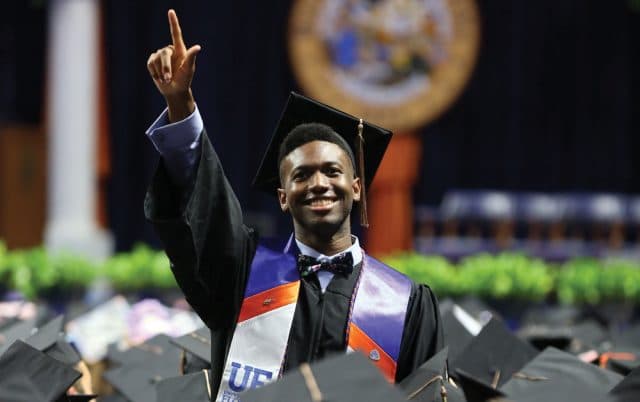 Student at graduation in regalia, pointing his hand to the sky and smiling