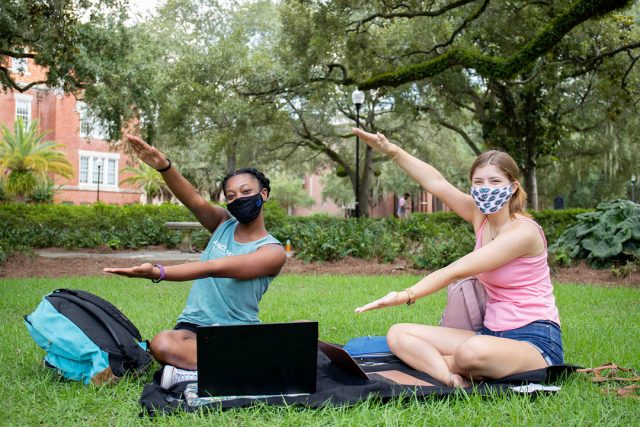 Two students sitting on the lawn, wearinging masks and doing the Gator Chomp