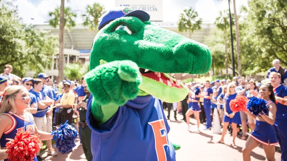 Albert Gator points into the camera while cheerleaders and fans assemble for a football game day
