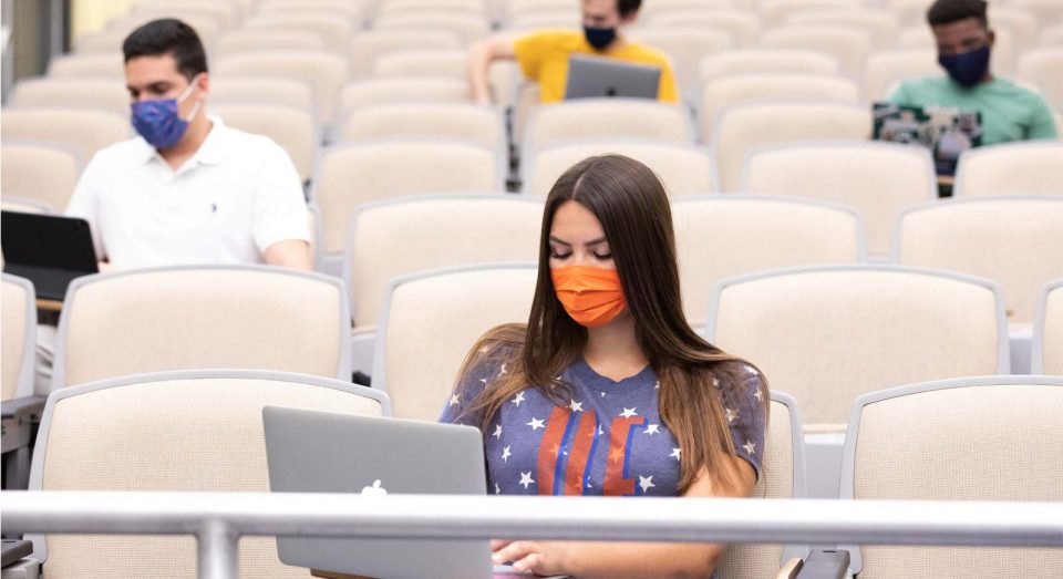 Students on laptops spread out among rows of seats with masks on
