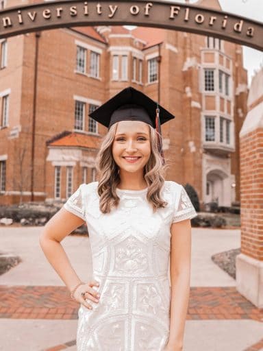 Alexa Entringer wearing her graduation cap under the University of Florida gateway with Heavener Hall in the background