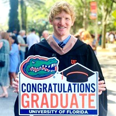 Charles (Wade) McCrea in his graduation gown holds a sign that says Congratulations Graduate, University of Florida