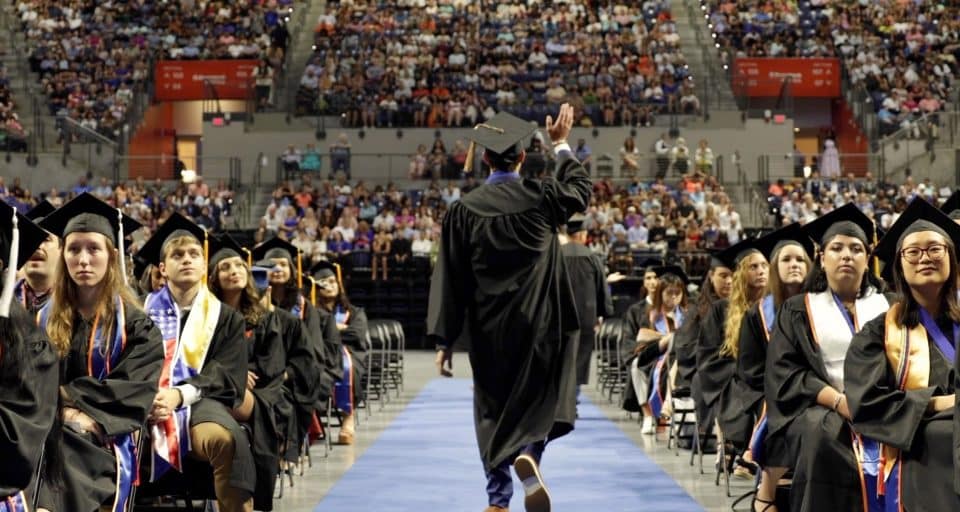 A graduate waves as he walks down the center of the O'Connell Center filled with graduates and other attendees