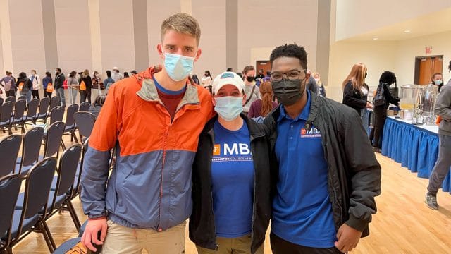 Three students with COVID masks on stand together for a photo while students behind them are in line to get refreshments