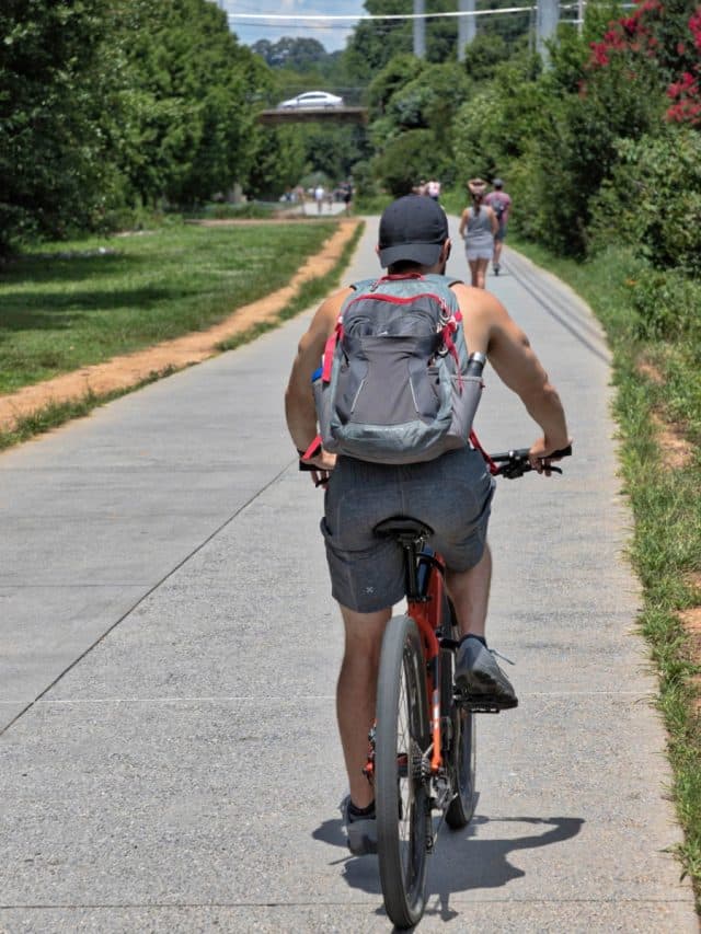 Multi-use paved path with a person on a bicycle, many people walking, and one on a scooter with grass and trees lining the path