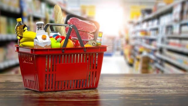 A basket of groceries sits on a table with a grocery aisle in the background