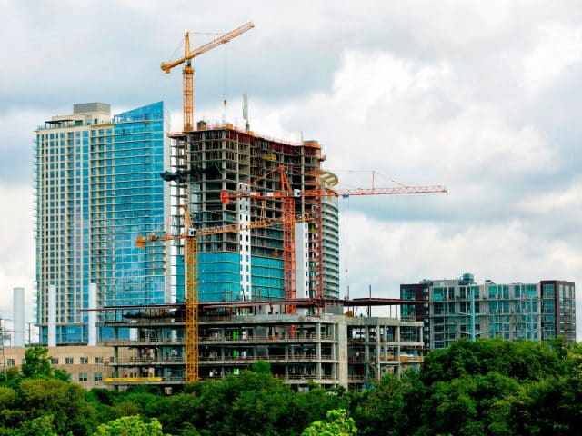 An apartment tower under construction in Austin, Texas with two cranes on site, buildings in the background and trees in the foreground