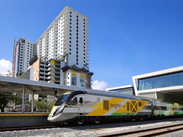 A Brightline train leaves West Palm Beach with the ParkLine tower in the background