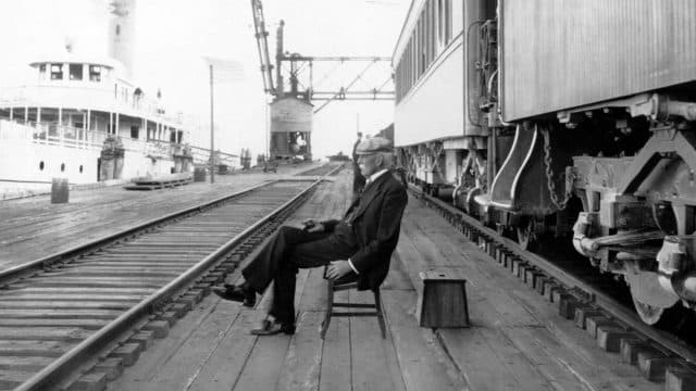 A black and white photo of Henry Flagler seated in a chair between railroad tracks outside his private rail car on Knights Key in 1908 with a ship docked nearby
