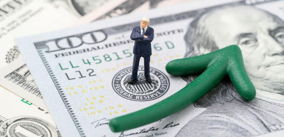 A small figure of a man stands on a 100 dollar bill with a green arrow curving upward