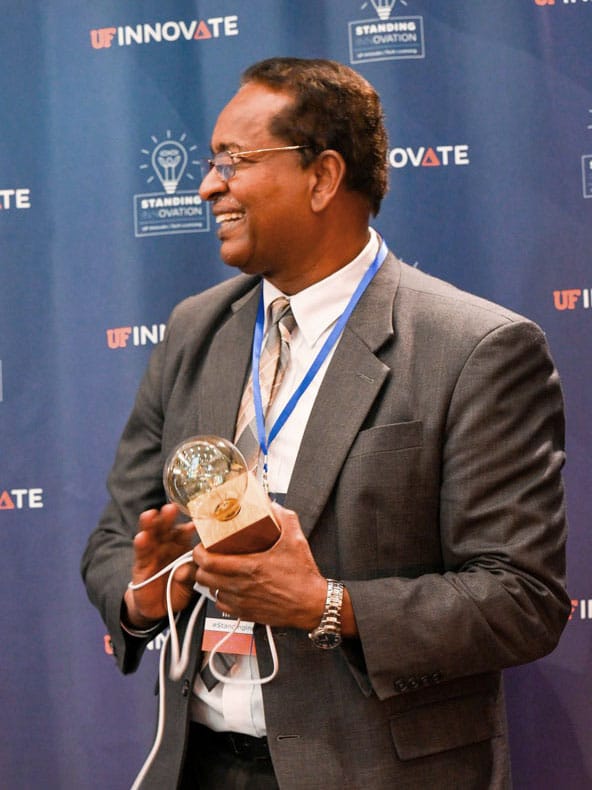 Nawari Nawari holding an award that is a lightbulb on a block of wood with a cord to plug it in and a UF Innovate background