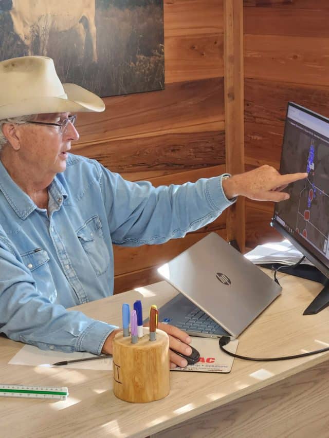 Jim Strickland points to a location on his computer screen as he wears a cowboy hat at a desk with a laptop and mouse