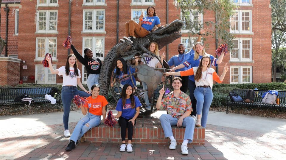 Students standing or sitting around, on or in the Gator Ubiquity Statue hold orange and blue pompoms, or a number one finger in the air