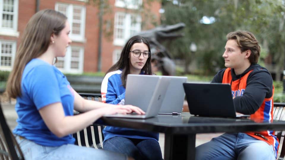 Three students with laptops sit at a table between Heavener Hall and Bryan Hall with the Gator Ubiquity Statue in the background