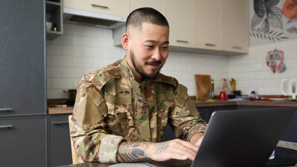 A man in military clothing works on a laptop at a kitchen table