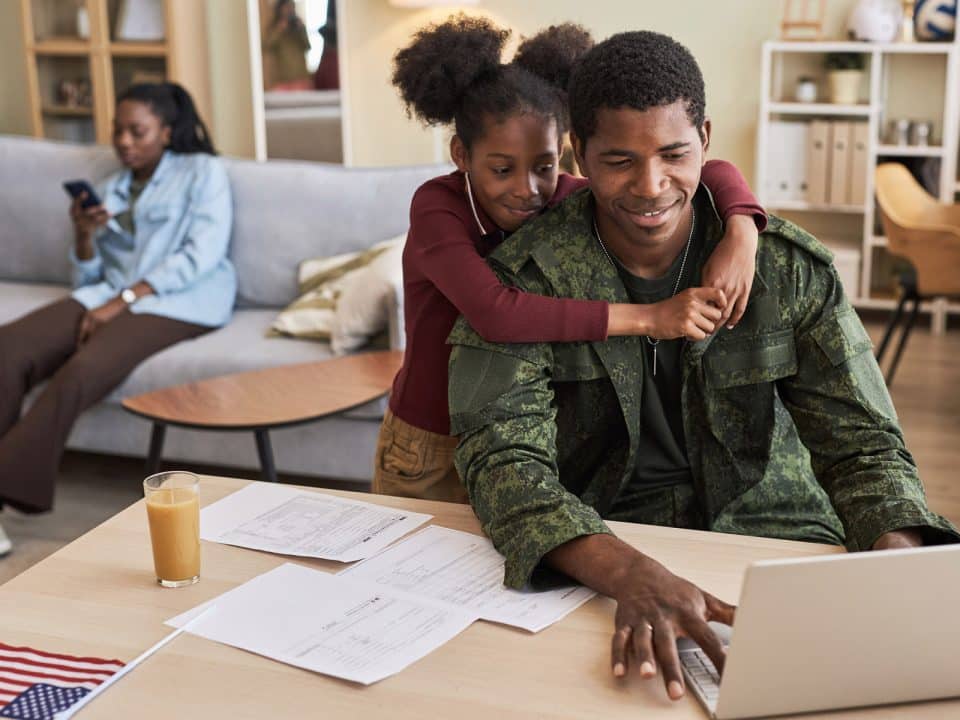 A man in military clothing works on his laptop in a living room while a young girl puts her arms around him and a woman on the couch in the background looks at her phone