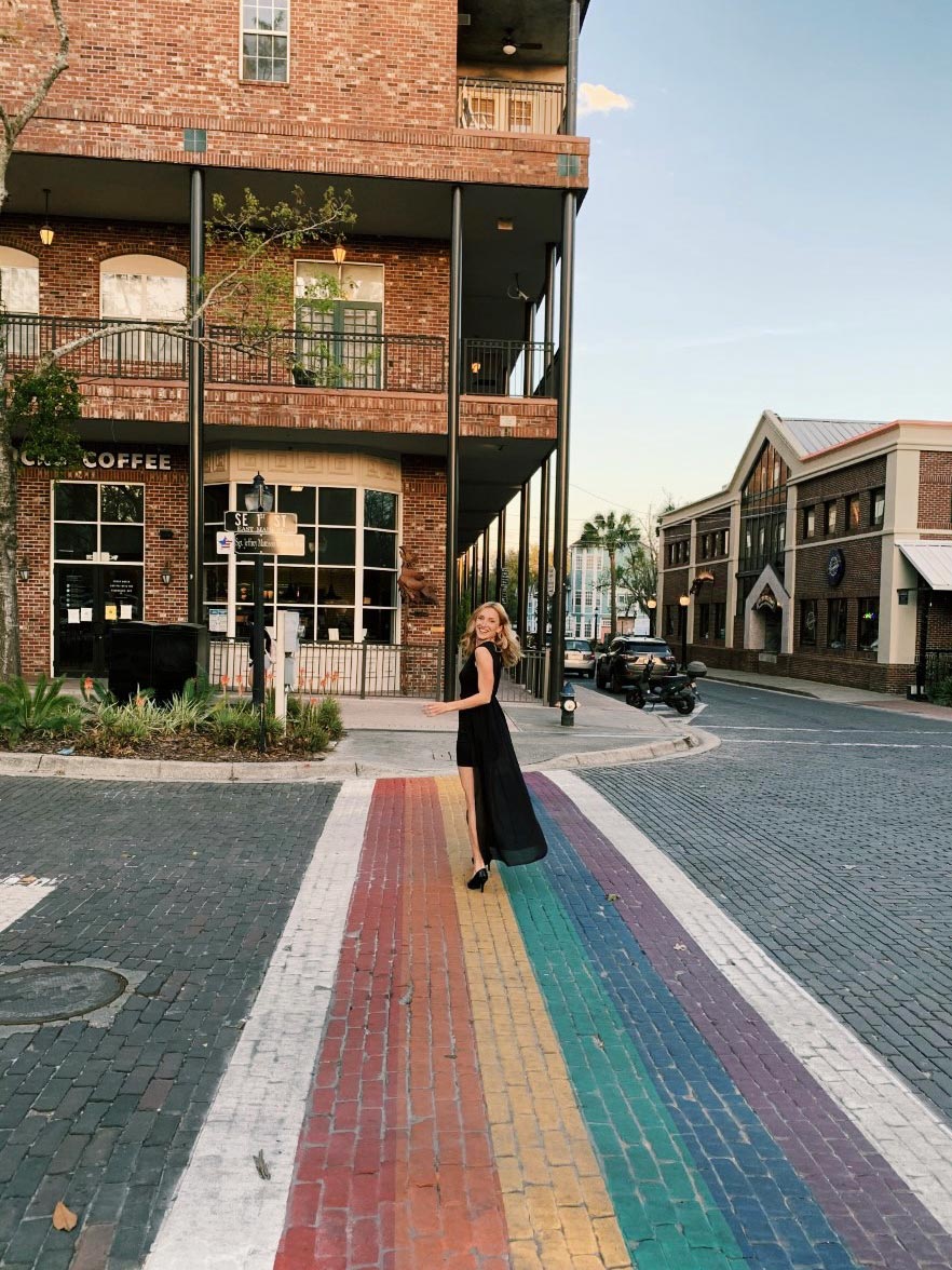 Person in an evening dress looks over her shoulder as she walks on the rainbow colored crosswalk, downtown Gainesville